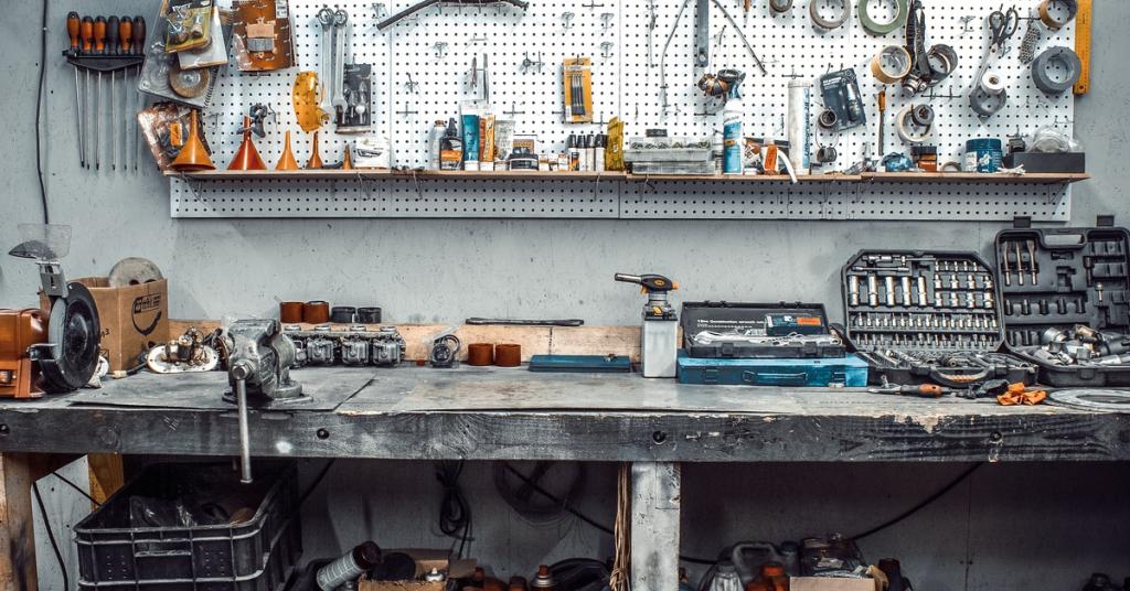 A worktable has many tools strew about. There are other tools attached to the wall by a peg board.