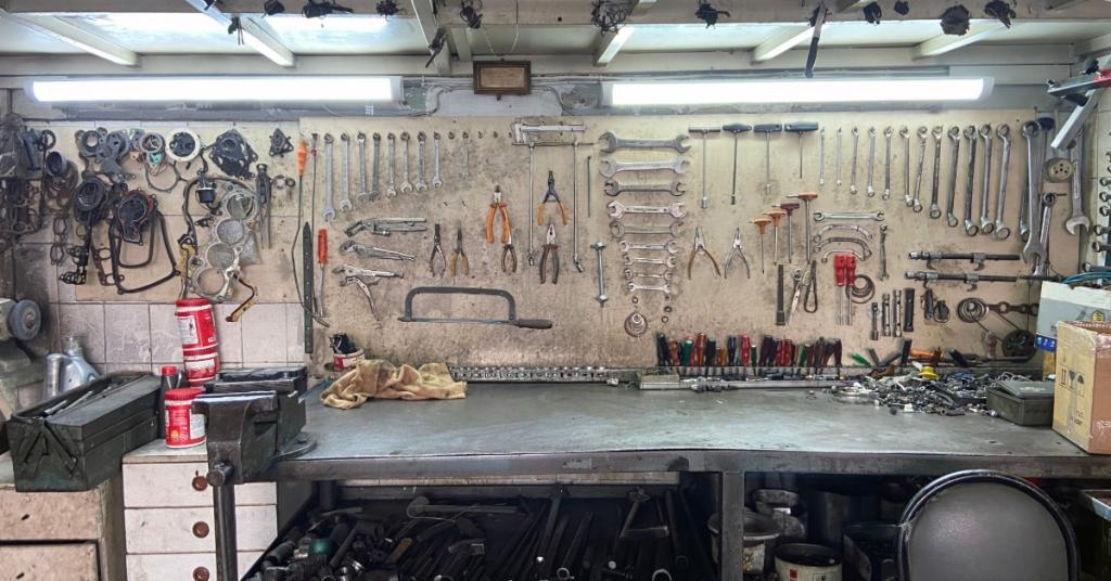 A workshop with a desk and a wall filled with tools. There are shelves under the work table.
