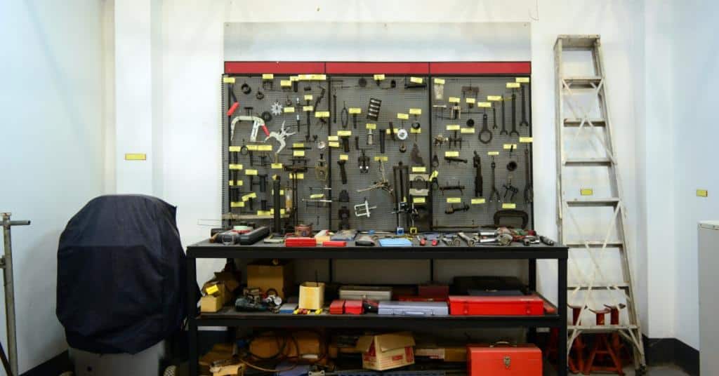 Tools are hanging on some wall storage. There is also a ladder to the side of the tool wall hangings.