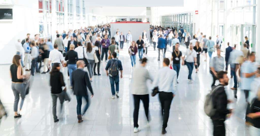 A large gathering of people are in a large, lobby-like area. There are window walls on the left and right of the room.