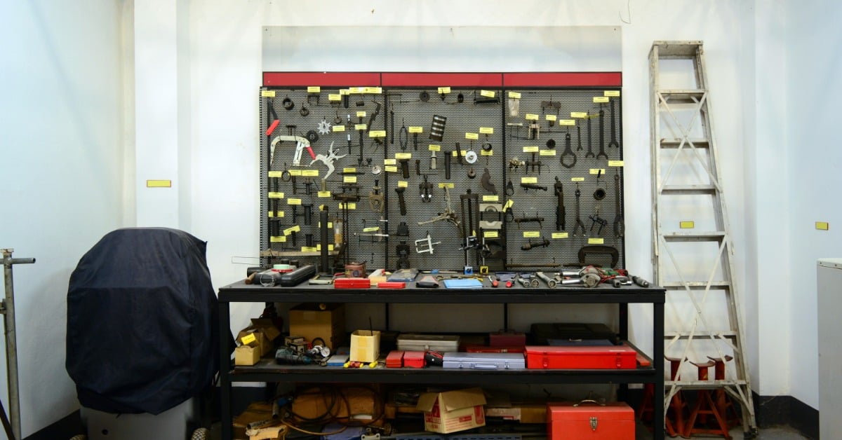 A workshop with a wall of tools hanging. There is a ladder to the right of the workshop bench.