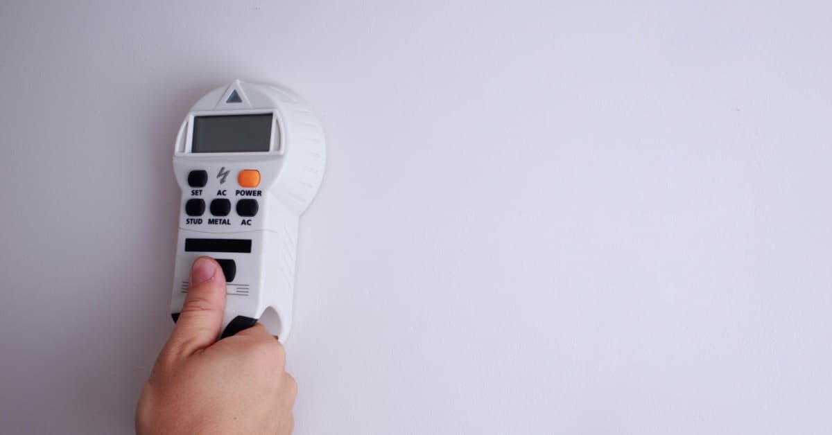 A person is using a stud finder against a wall. The wall is white in color and has no other adornments.