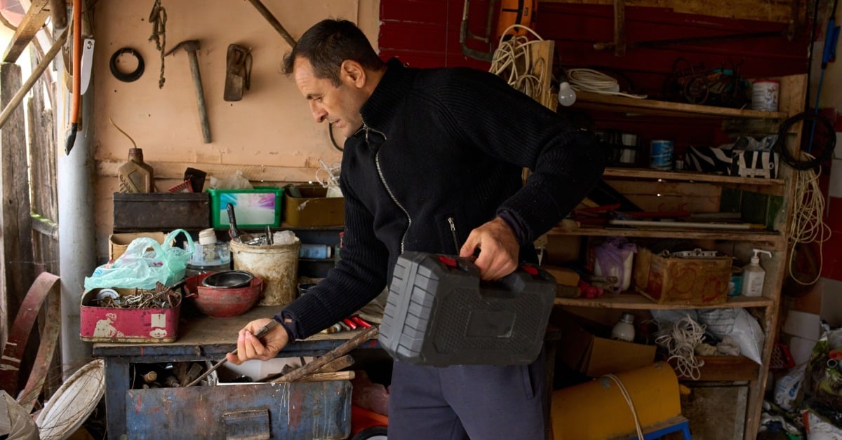 A man is standing in a garage and is looking through drawers. He has a tool box in his free hand.
